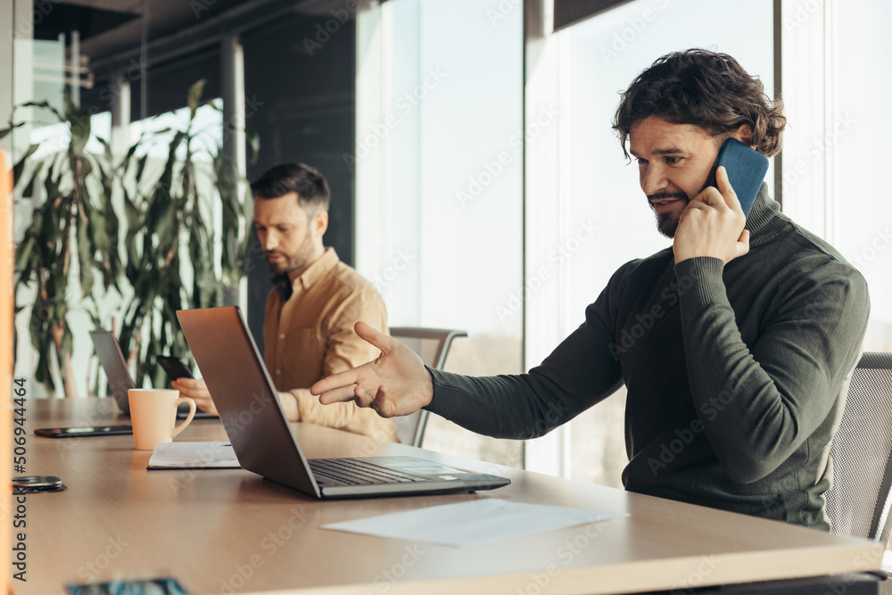 Handsome young businessman using laptop, speaking on smartphone, working with male colleague at modern office Handsome young businessman using laptop, speaking on smartphone, working with male colleague at modern office
