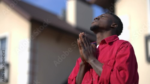 Religious African American young man with clasped hands praying standing in sunlight outdoors. Hoping handsome guy thanking God enjoying sunny calm day on backyard