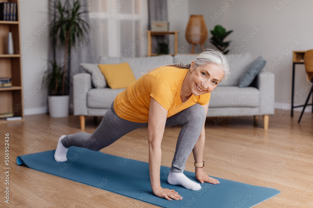 Athletic aged woman stretching her legs, doing runner's lunge yoga pose on home workout, exercising in living room