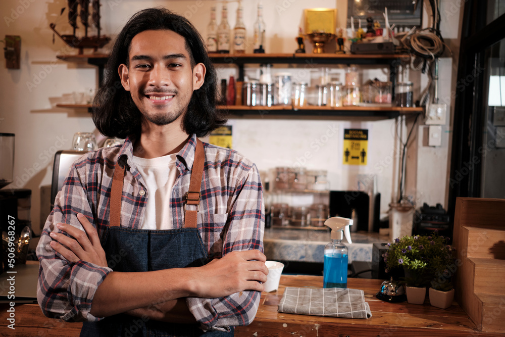 Long hair handsome Asian male startup barista with apron stands at casual cafe counter bar, arms crossed, looks at camera with and welcoming smile, happy and cheerful with coffee shop service jobs.