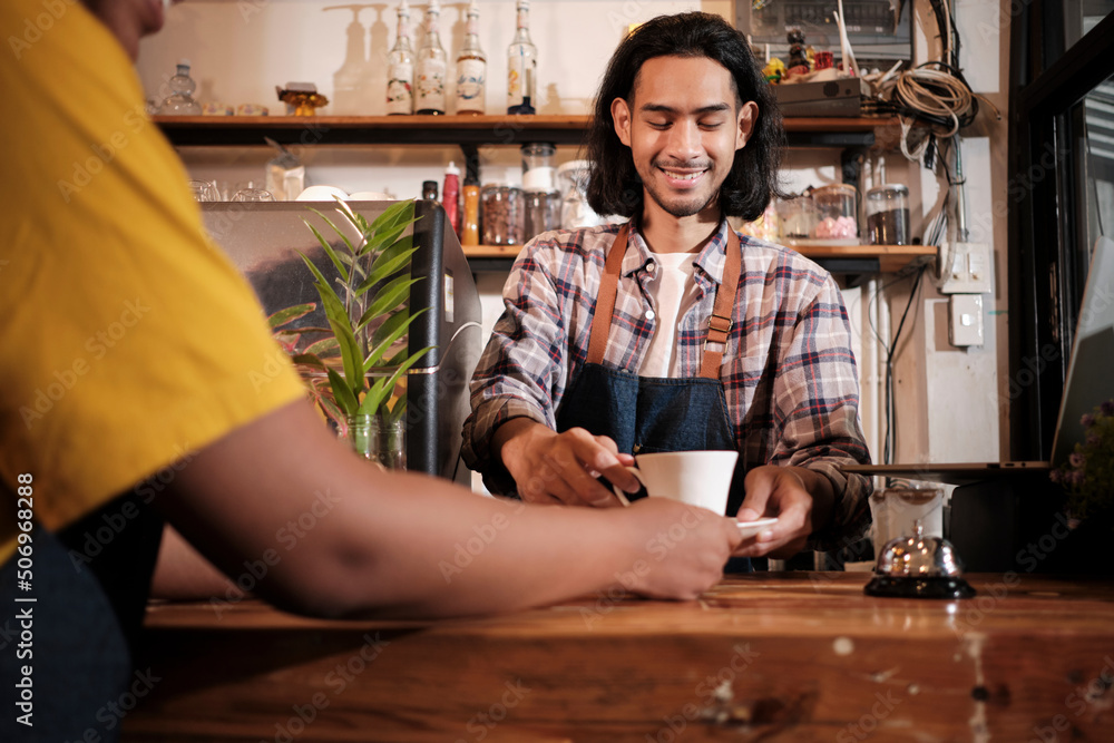 Close-up shot, young long hair Thai male baristas startup works by serving a cup of coffee to a customer with a smile at casual cafe counter bar, happy service job, and small business entrepreneur. Close-up shot, young long hair Thai male baristas startup works by serving a cup of coffee to a customer with a smile at casual cafe counter bar, happy service job, and small business entrepreneur.