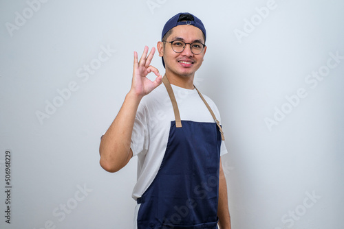 Young handsome barista man wearing apron smiling and doing ok sign with fingers, excellent symbol