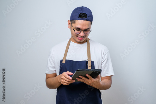 Cheerful young Asian barista man wearing apron and cap using digital tablet to manage cafe order