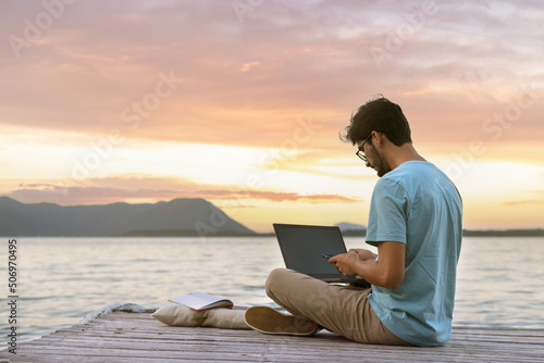 Young digital nomad man sitting on wooden pier at sea working on internet remotely at sunset - Traveling with a computer - Online dream job concept. photo