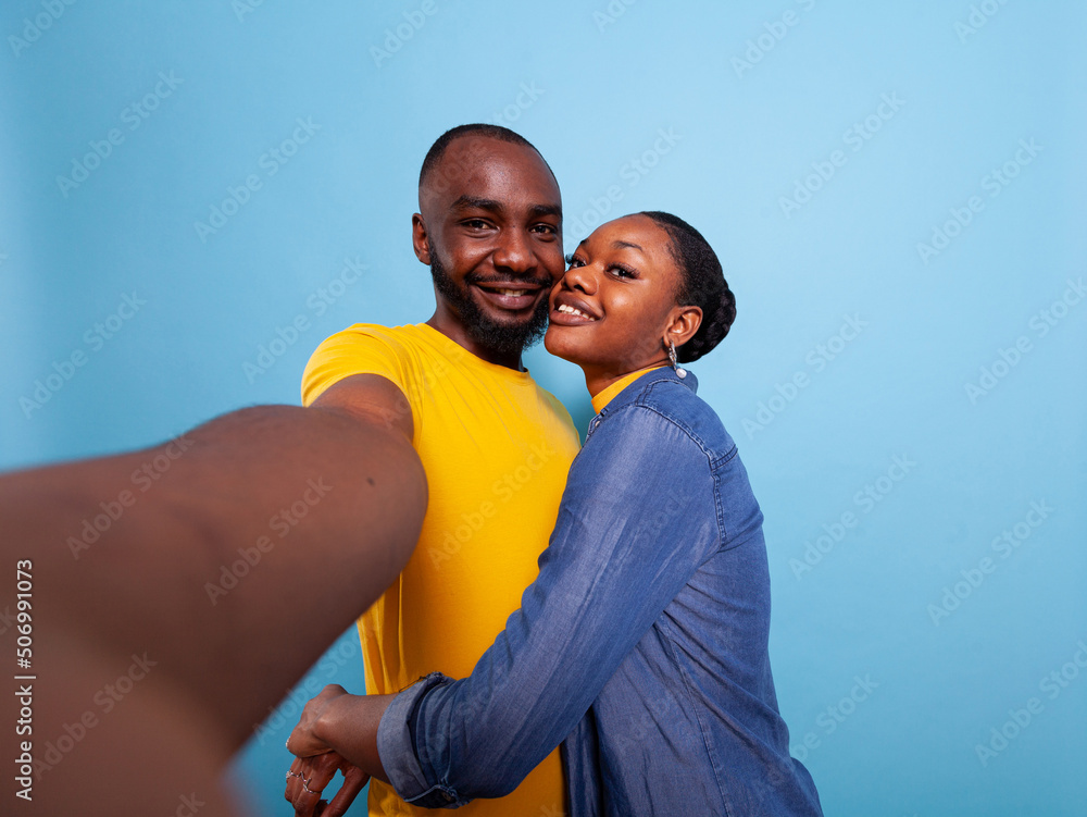 Obraz premium POV of man holding camera and woman hugging him in studio, taking pictures. Romantic couple sharing affection and love together, embracing over blue background. Happy people in relationship.
