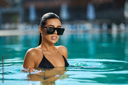 Sexy brunette woman wearing black bikini relaxing in swimming pool. Front view of young lady in swimwear and sunglasses looking at camera, while swimming in pool in luxury resort. Concept of leisure.