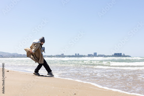 a six-year-old girl in a beige trench coat touches the water with her foot