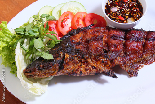 Patin fish (siamese sha (siamese shark, sutchi catfish), which is grilled and grilled whole. With a complement of lettuce, cabbage, basil and tomatoes. Plus spicy soy sauce. White plate. Wooden table. photo