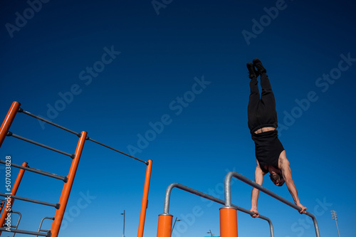 Shirtless man doing handstand on parallel bars at sports ground. 