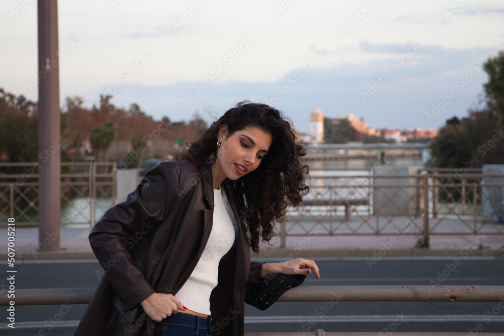 Fototapeta premium Young and beautiful woman with dark and curly hair is sightseeing in Seville. In the background part of the Mediterranean style European city in the golden hour. Tourism and holidays concept.