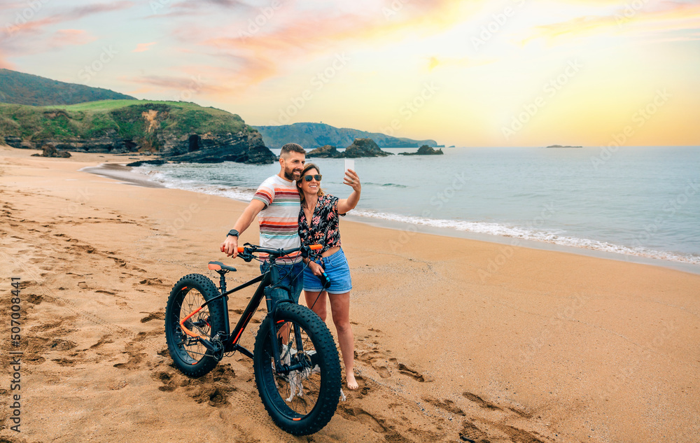 Fototapeta premium Young couple with fat bike taking a selfie with the mobile on the beach