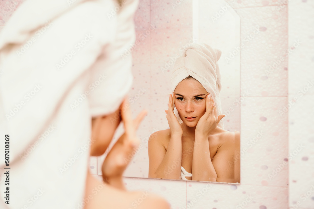 Young beautiful woman using face cream moisturizing lotion after bath. Pretty attractive girl in a towel on her head stands in front of a mirror in a home bathroom. Daily hygiene and skin care Young beautiful woman using face cream moisturizing lotion after bath. Pretty attractive girl in a towel on her head stands in front of a mirror in a home bathroom. Daily hygiene and skin care
