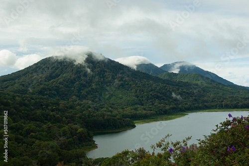 Highlands landscape on Bali - mountains with lush green forest, lake and little flowers in sunny and cloudy day with fluffy white clouds on peaks, panorama. Amazing travel on Indonesia and asia.