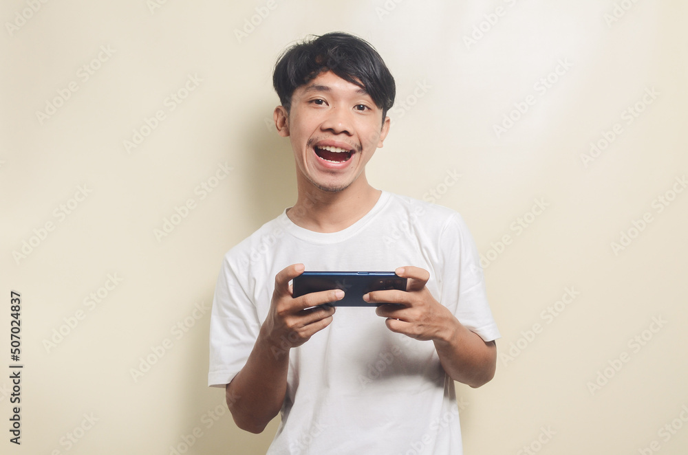 asian man wearing white t-shirt with gesture playing game on smartphone, isolated background asian man wearing white t-shirt with gesture playing game on smartphone, isolated background