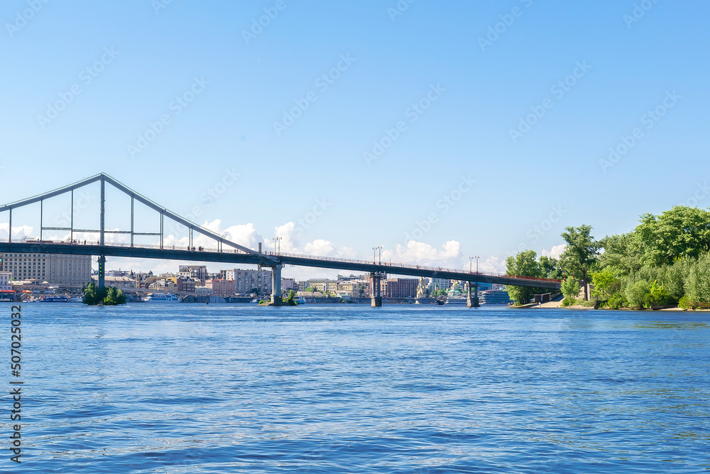 Scenic nature travel cityscape river Dnipro with blue water and metal pedestrian bridge at sunny summer day in Kiev. View from inside river, ship on water Scenic nature travel cityscape river Dnipro with blue water and metal pedestrian bridge at sunny summer day in Kiev. View from inside river, ship on water