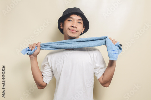 happy asian man wearing bright white t-shirt with black hat and carrying towel on isolated background