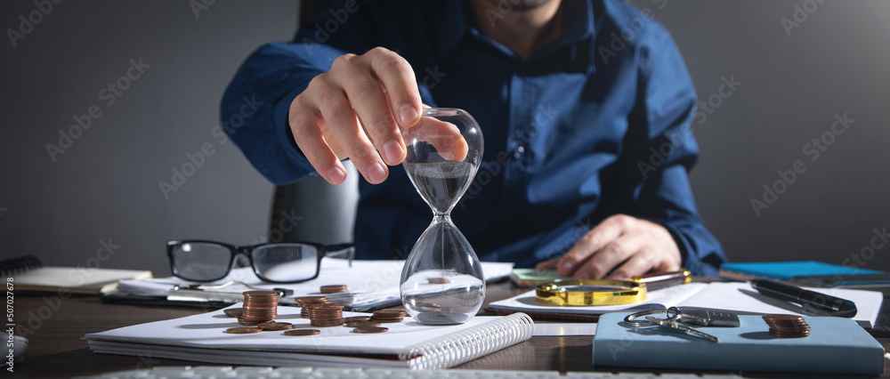 Fototapeta premium Businessman holding hourglass. Coins and business objects on the desk