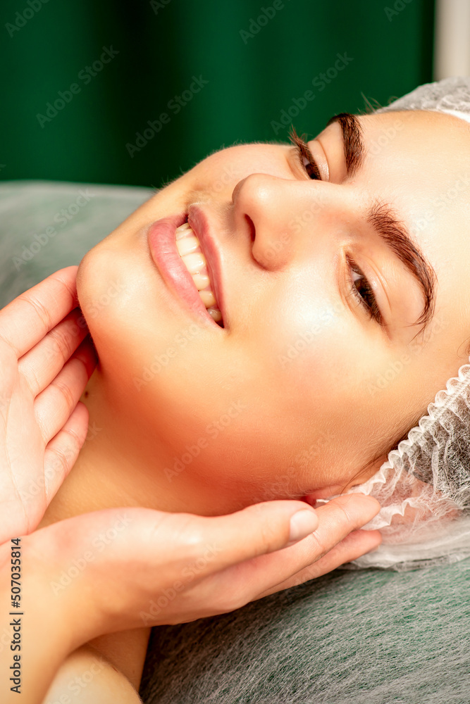 Naklejka premium Plastic surgery and beauty concept. Professional cosmetician examining and touching the face of the beautiful young caucasian smiling woman in a clinic of esthetic cosmetology