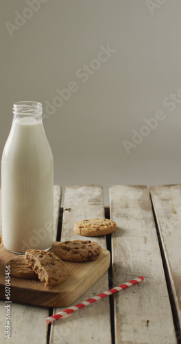 Vertical image of chocolate chip cookies and bottle of milk on chopping board and wooden background