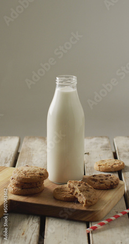 Vertical image of chocolate chip cookies and bottle of milk on chopping board and wooden background