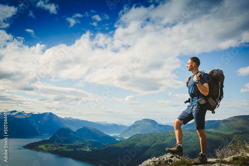 Active hiker with backpack standing on a rocky mountain ridge looking out of green alpine valley and peaks. Healthy outdoor lifestyle concept