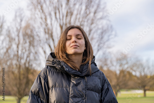 Young teen girl with closed eyes standing outdoors in park under sunshine