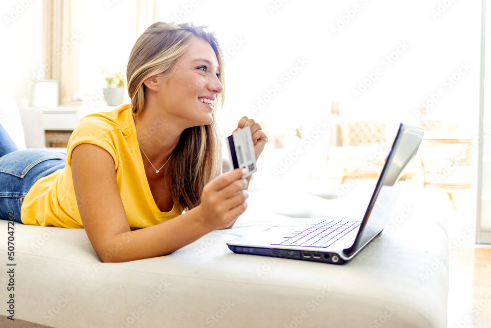 Cheerful woman doing online shopping while lying on a couch at home. Young girl holding her credit card while using her laptop. People, internet bank, online shopping, technology and e-money concept. Cheerful woman doing online shopping while lying on a couch at home. Young girl holding her credit card while using her laptop. People, internet bank, online shopping, technology and e-money concept.