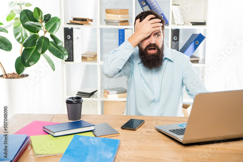 Frustated man clutching head working at laptop in office, frustration photo
