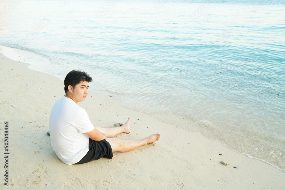 Man wearing white t-shirt and black short sitting on sand at the beach in morning light. Looking far away on sea water. Feeling thoughtful, calm, relax, loneliness, silent with myself.