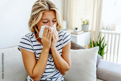 Shot of an attractive young woman feeling ill and blowing her nose with a tissue at home. Photo of sneezing woman in paper tissue. Picture showing woman sneezing on tissue on couch in the living-room.
