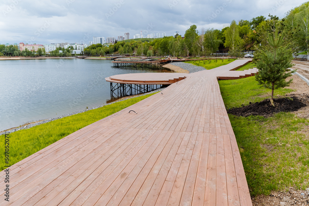 A wooden road along the embankment of the lake, the construction of a city park on a pond, a deck of boards, modern landscape design, landscaping of the city, a place to relax by the water.