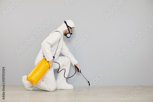 Pest control guy cleaning home from insects. Man in white protective suit crouching near wall and spraying floor with cockroach insecticide from yellow bottle for safe living environment inside house photo