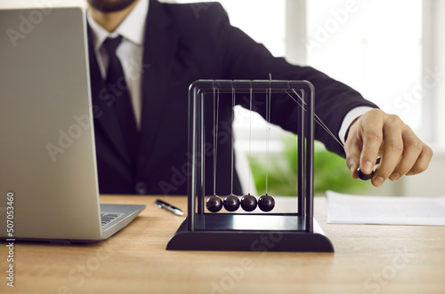 Close up of business leader playing with Newton's cradle on office desk with notebook PC. Man moves black pendulum ball, it hits others, and kinetic energy is transferred into potential and vice versa