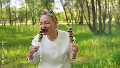 Mature woman 50 years old eating grilled vegetables from wooden skewers outdoors in forest 