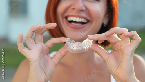 Red-haired Caucasian woman holding transparent mouthguards for bite correction outdoors. A girl with a beautiful snow-white smile uses silicone braces