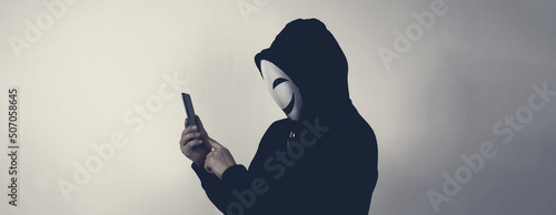 Anonymous hacker and face mask with smartphone in hand. Man in black hood shirt holding and using mobile phone on white background. Represent cyber crime data hacking or stealing personal data concept photo