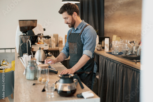 Handsome barista wearing apron uses a digital tablet to takes order from clients at coffeehouse