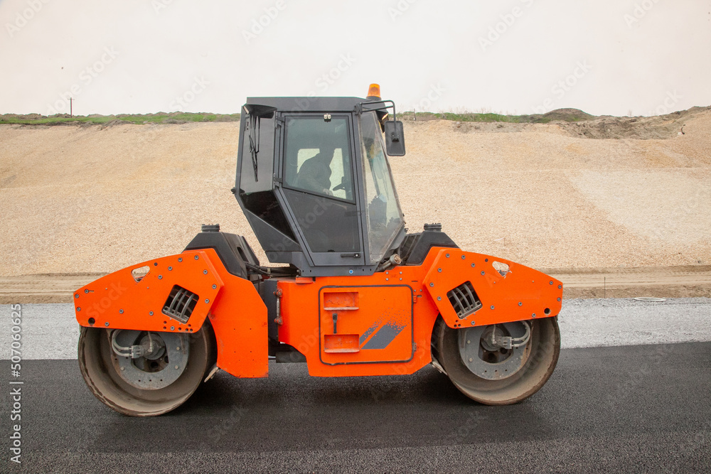 An attractive new orange road roller with a climate controlled enclosed cab sits on an unfinished new road. An attractive new orange road roller with a climate controlled enclosed cab sits on an unfinished new road.