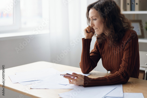 Thoughtful serious unhappy curly cute lady sitting at table with papers documents worried about problem at work think about failure bankruptcy calculates budget looks aside. Money problem concept