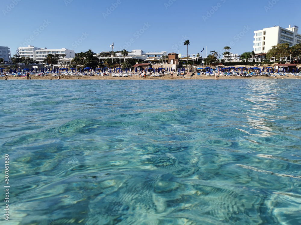 Protaras. Famagusta area. Cyprus. View from the sea to the beach of Fig Tree Bay, where people sunbathe and swim, to hotels and restaurants against a cloudless sky.