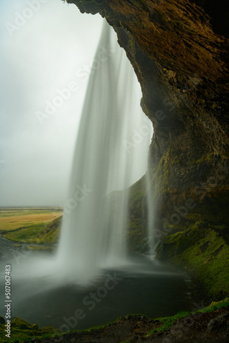 Long exposure behind an Icelandic landmark waterfall in greenish tones  dark lake  framed by yellow cliff and monotonous grey sky