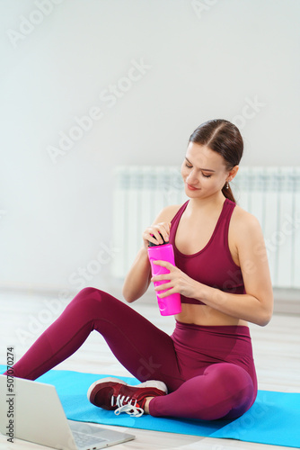 Fototapeta Naklejka Na Ścianę i Meble -  a young woman drinks water in a burgundy tracksuit with laptop on a mat in a gym