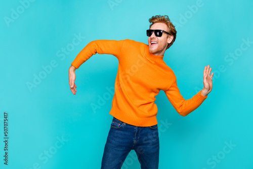 Photo of funky sweet young man wear orange pullover dark spectacles dancing smiling isolated teal color background