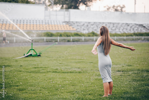 A girl in a gray dress stands with her hands apart and waits. lawn sprinkler sprays water in stadium © maxfotoadobe