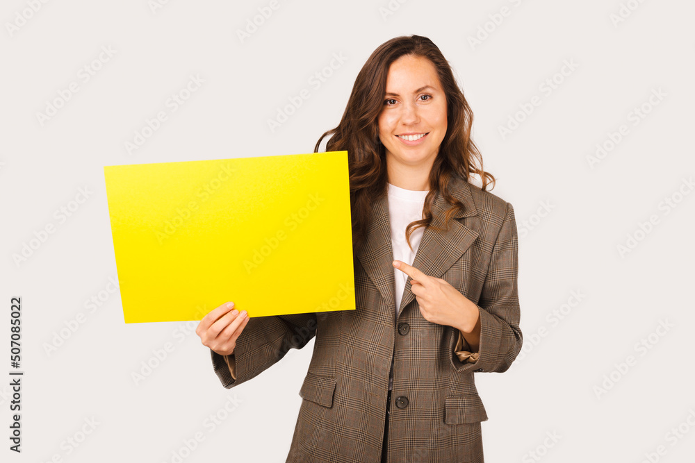 Young woman is holding a yellow placard with copy space and pointing at it. Studio shot over white background. Young woman is holding a yellow placard with copy space and pointing at it. Studio shot over white background.