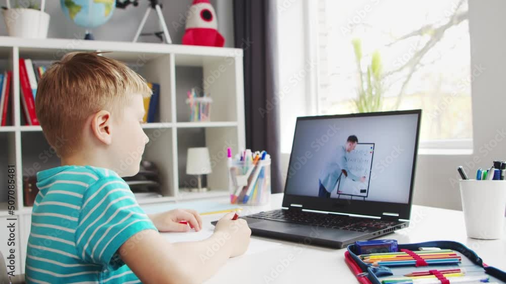 Boy is Doing his Homework at the Table. Cute Child is Learning at Home with the Help of Laptop Computer and School Textbooks. Study and Distant Education Concept. Boy is Doing his Homework at the Table. Cute Child is Learning at Home with the Help of Laptop Computer and School Textbooks. Study and Distant Education Concept.