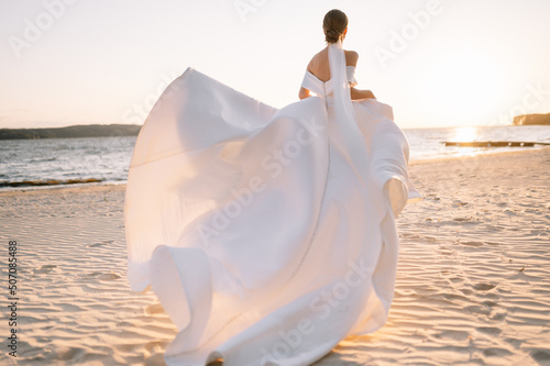 Happy young bride woman in white dress running, have fun on clean sandy beach waves of the sea or ocean on sunset, summer photo shooting near a water. Wedding rest, relax honeymoon concept.