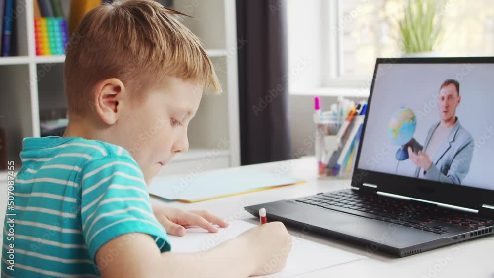 Boy is Doing his Homework at the Table. Cute Child is Learning at Home with the Help of Laptop Computer and School Textbooks. Study and Distant Education Concept. Boy is Doing his Homework at the Table. Cute Child is Learning at Home with the Help of Laptop Computer and School Textbooks. Study and Distant Education Concept.