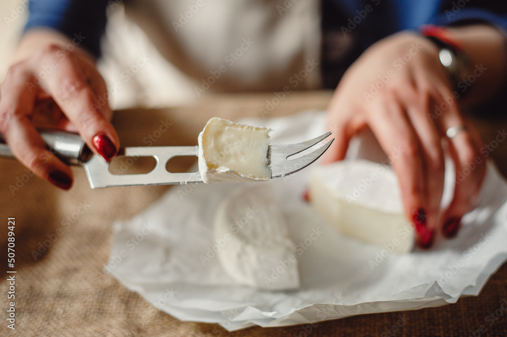 Obraz premium Cut Camembert cheese, French soft cheese with white mold in the hands of a woman on the background of a craft textile tablecloth, close-up. Knife for slicing cheese