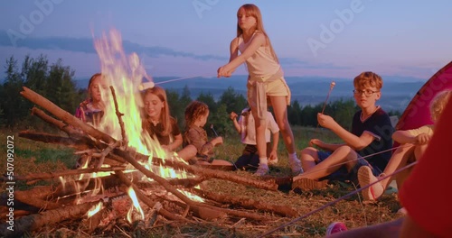 Side view of group of kids sitting around big campfire with roasted meal at twilight. Happy teenagers frying sausages and marshmallow over fire, with panoramic view on background. Concept of camping.